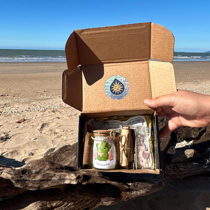 The Heart Moonling package opened with contents on a beach showing the glass jar with a cork lid, the Heart Moonling sticker, hangtag, and scroll