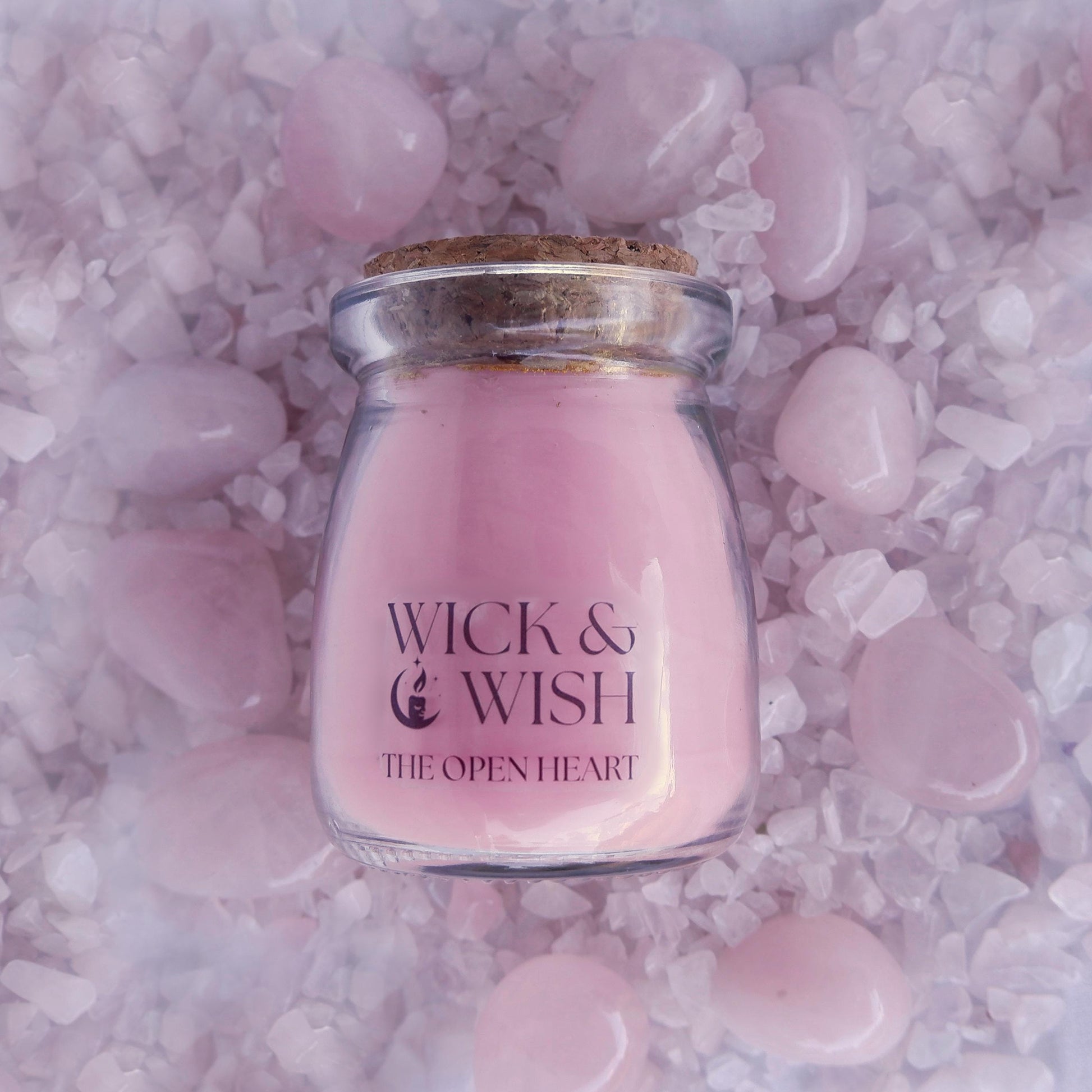 The Open Heart Candle in a glass jar with cork lid on pink rose quartz stones, featuring 'Wick & Wish' branding.