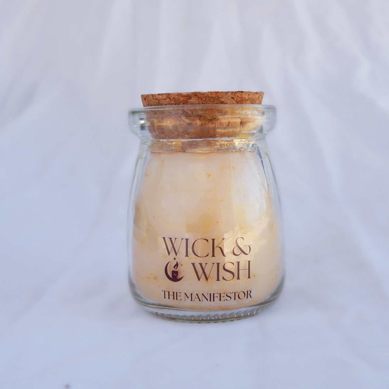 Candle in a glass jar with cork lid on a white background, featuring 'Wick & Wish The Manifestor' branding.