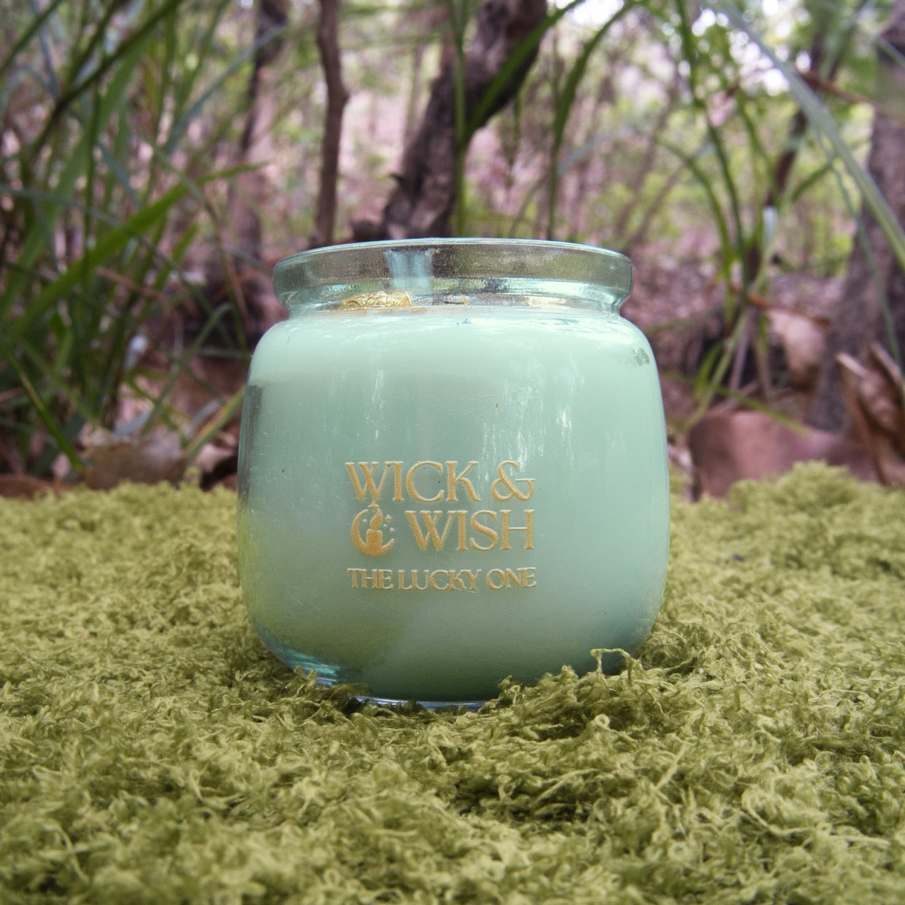Green Candle jar label ed 'Wick & Wish The Lucky One' on a grassy surface with a natural background