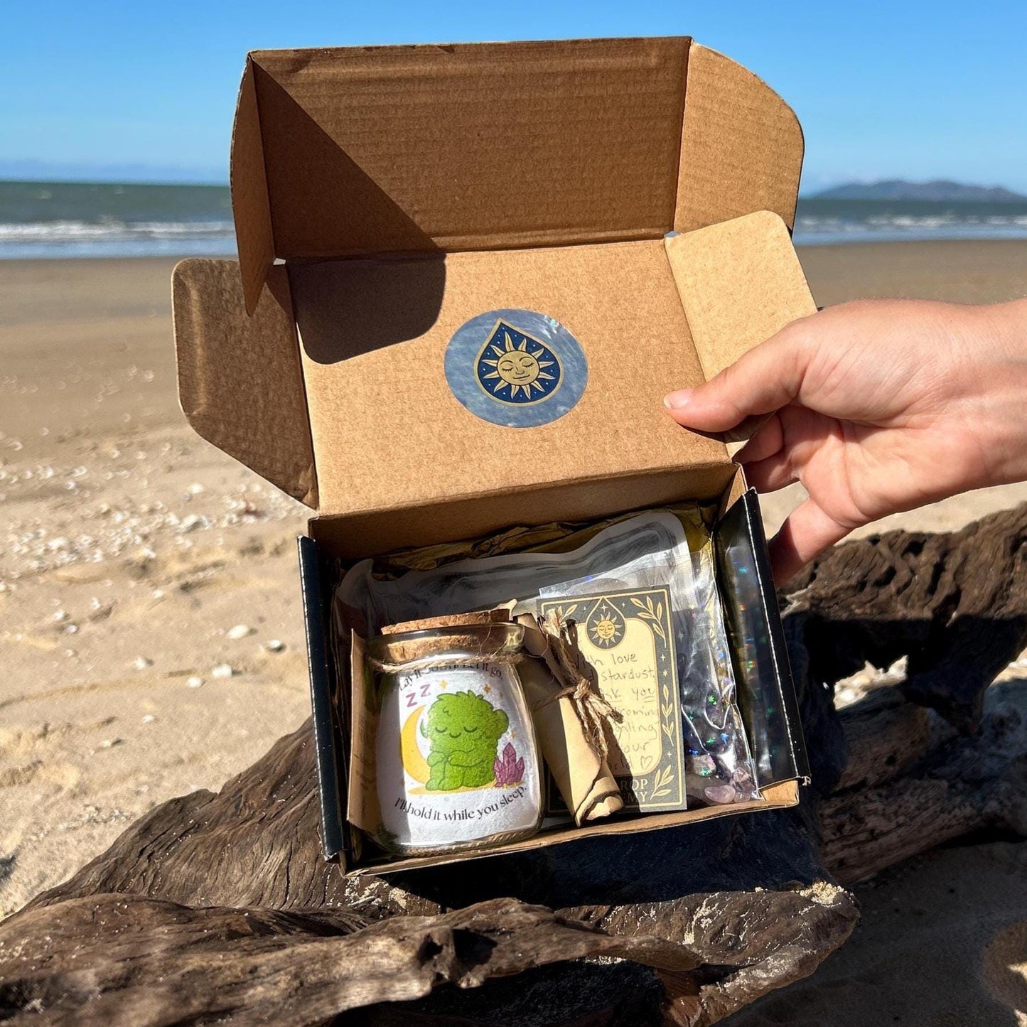 Amethyst Dream Moonling box opened on the beach showing a glass jar with a cork, a Dream Moonling cartoon sticer, scroll, and hangtag visible